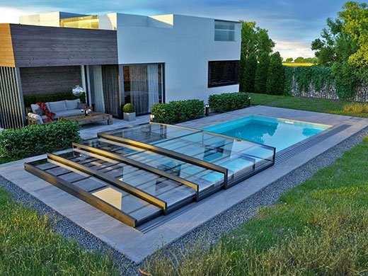 Modernes Haus mit Schiebedach-Pool, graue Fassade, große Fenster, grüne Umgebung.