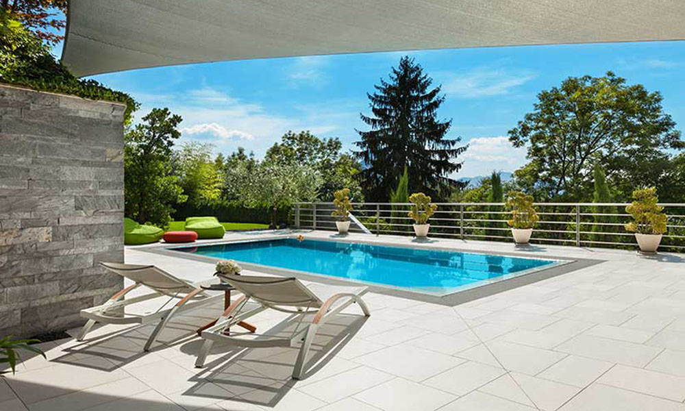 Zwei weiße Liegestühle am Pool in einem modernen Garten, sonnig, mit grünen Pflanzen und Bäumen.