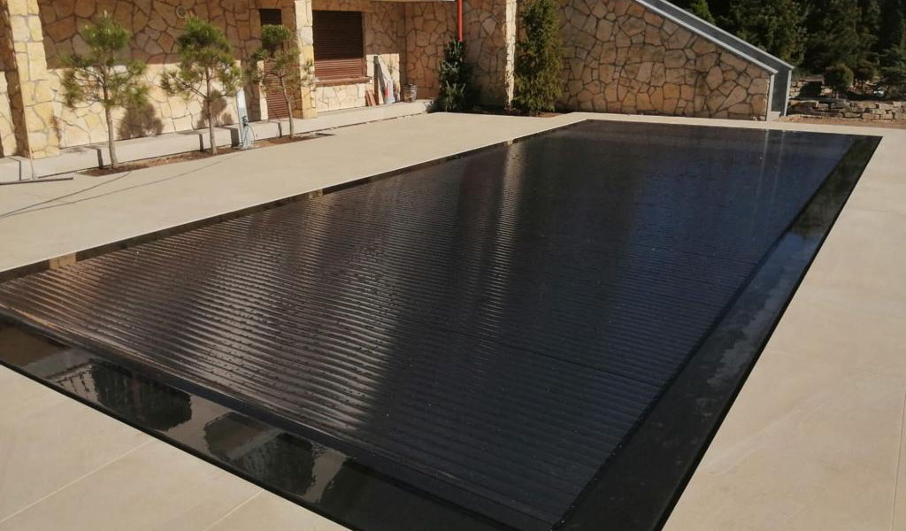 Ein moderner, rechteckiger Pool mit schwarzer Abdeckung auf einem Steinboden vor einem Haus.