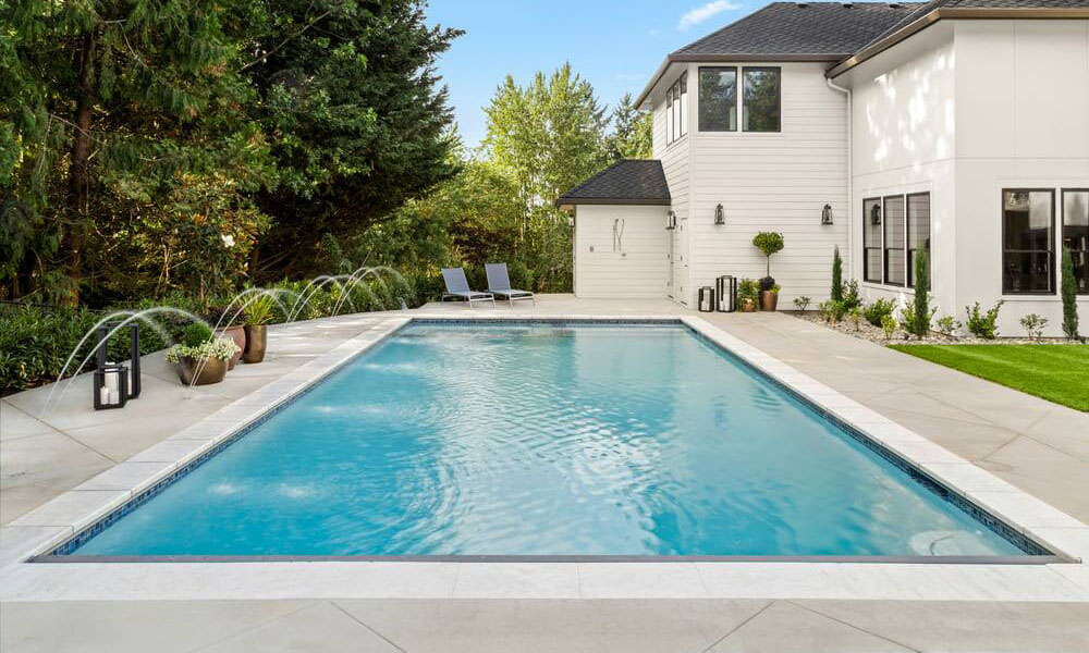 Großer Swimmingpool vor einem modernen, weißen Haus, umgeben von Bäumen und zwei Liegestühlen.