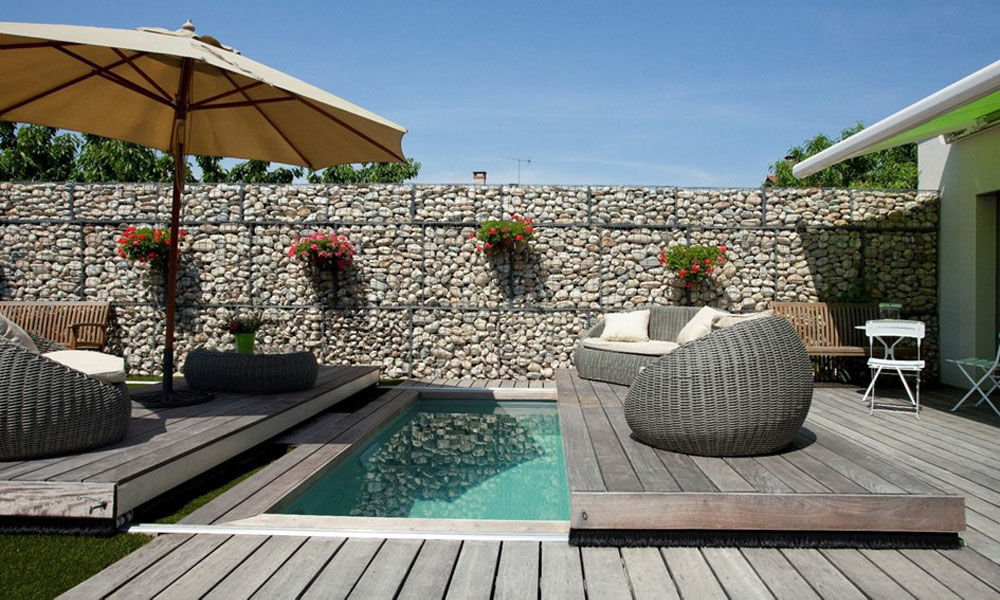 Deck mit kleinem Pool, Steinwand, Sonnenschirm, Rattanmöbel und Pflanzen; sonniges Wetter.