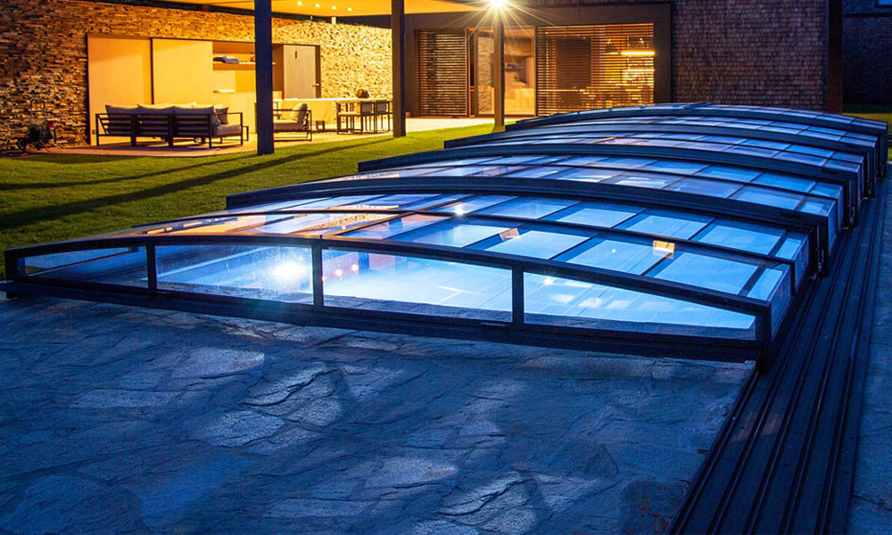Beleuchteter Pool mit Glasschutz in modernem Garten, Haus im Hintergrund, abends.